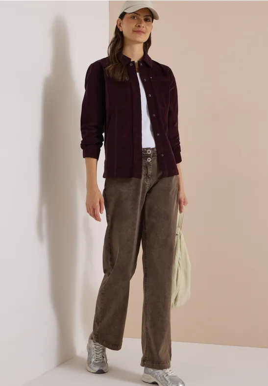 Frau im lässigen Look: weißes Top, bordeauxfarbene Jacke, braune Cordhose, silberne Sneaker, Cap.