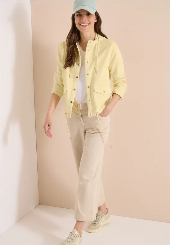 Frau steht in hellem Raum, trägt gelbe Jacke, weißes Top, beige Hose und Sneaker.