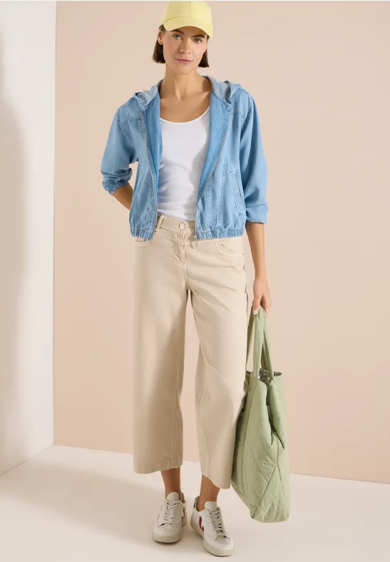 Eine Frau trägt ein weißes T-Shirt, eine hellblaue Jacke, beige Culottes, weiße Sneaker und eine grüne Tasche.