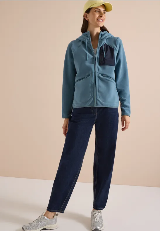 Frau in blauer Fleecejacke, dunklen Hosen, heller Cap und Sneakern, steht lachend vor neutralem Hintergrund.