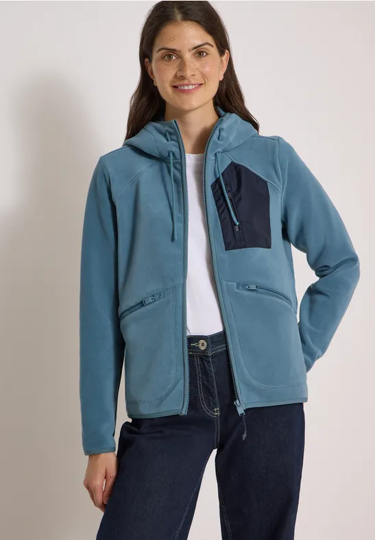 Eine lächelnde Frau trägt eine blaue Kapuzenjacke mit Reißverschluss und Brusttasche sowie eine Jeans.