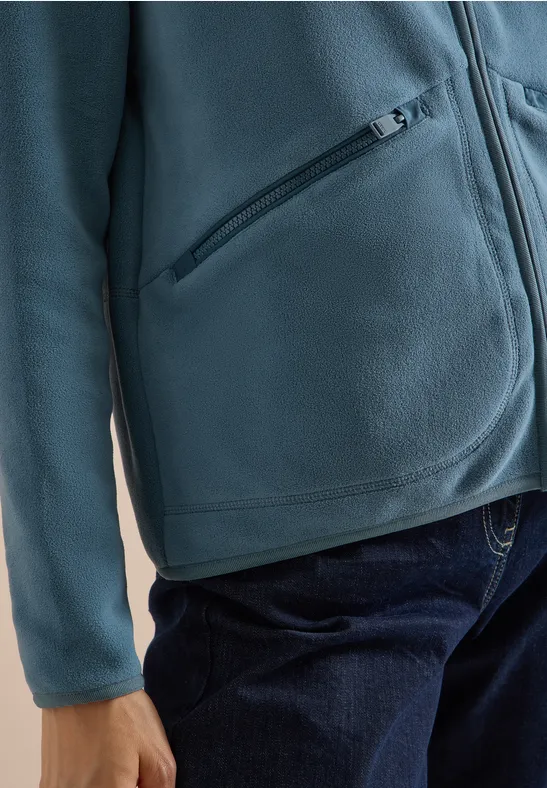 Person in türkisfarbener Fleecejacke mit Reißverschlusstasche und blauen Jeans.