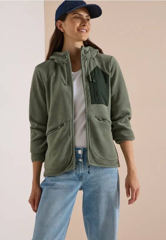 Frau trägt grüne Fleecejacke mit Kapuze, weiße Bluse, Jeans und Baseballcap.