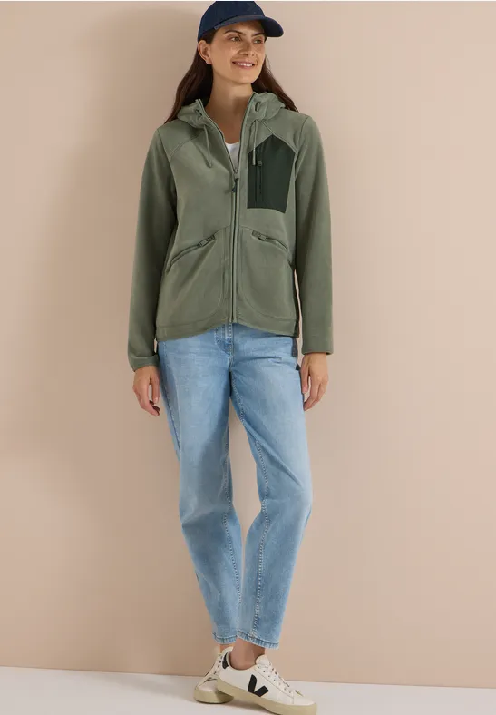 Frau in grüner Jacke, Jeans, Sneakern und Cap vor beigem Hintergrund.