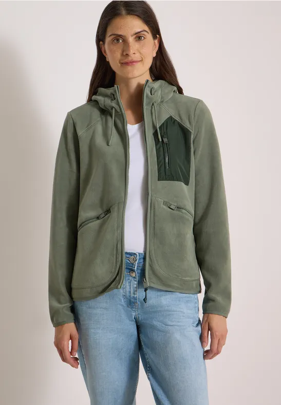 Eine Frau steht frontal, trägt eine grüne Fleece-Jacke mit Kapuze, weißes Shirt und hellblaue Jeans.