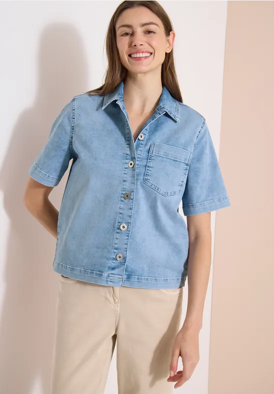 Frau in hellblauer Jeansbluse mit Knöpfen trägt beige Hose vor neutralem Hintergrund.