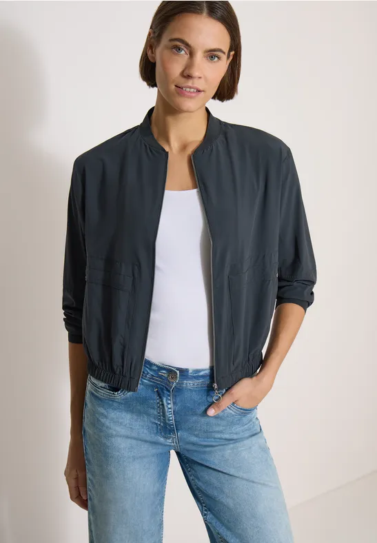 Frau trägt dunkle Bomberjacke, weißes Top und blaue Jeans.