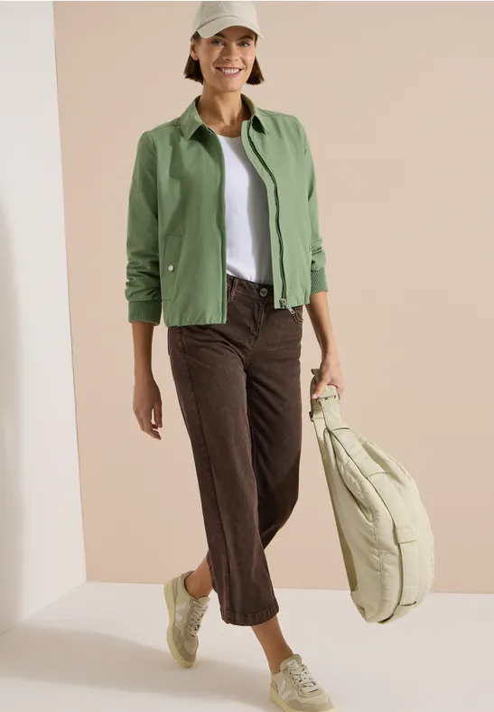 Frau trägt grüne Jacke, weißes Top, braune Cropped-Hose, grüne Sneaker und große beige Tasche.