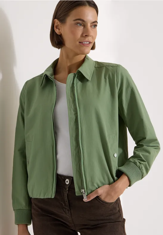 Person trägt grüne Hemdjacke, weißes Top und braune Hose in schlichtem, modischem Look.