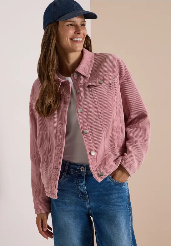 Junge Frau in rosafarbener Cordjacke, Jeans und blauer Cap lächelt entspannt.