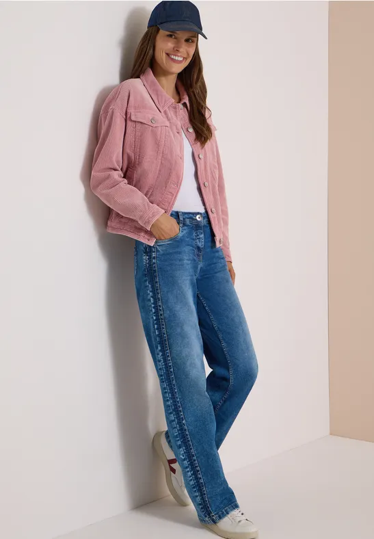 Eine lächelnde Frau mit rosa Cordjacke, Jeans und Cap lehnt lässig an einer Wand.