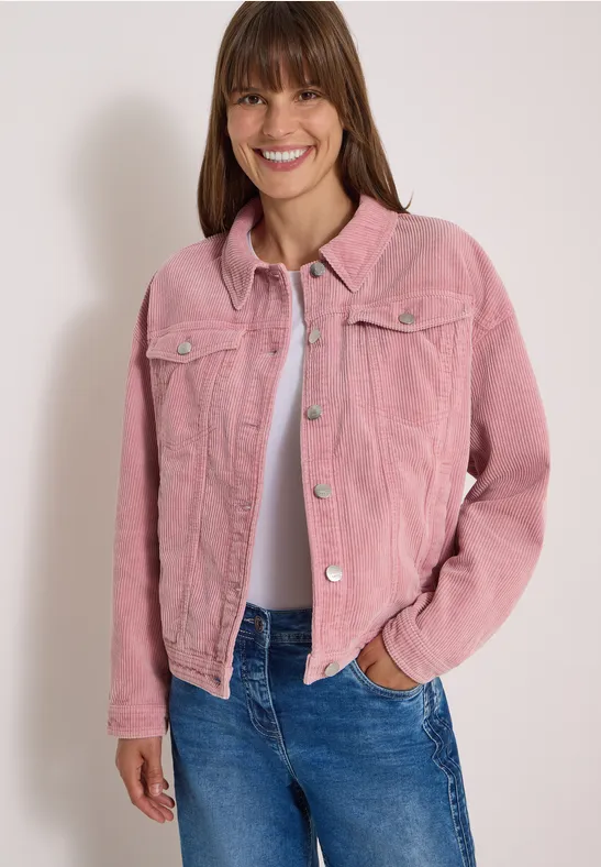 Fröhliche Frau in rosa Cord-Jacke, weißem T-Shirt und blauen Jeans vor neutralem Hintergrund.