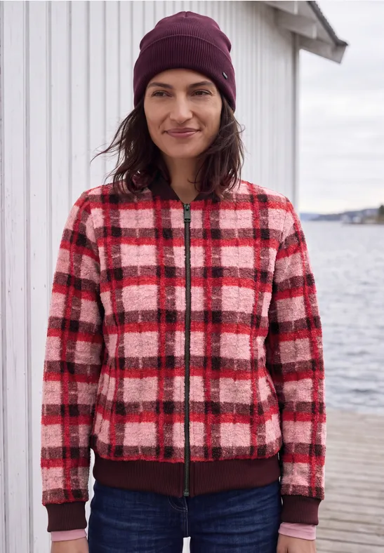 Femme en veste à carreaux rouge et blanche et bonnet, debout sur une jetée au bord de l'eau.