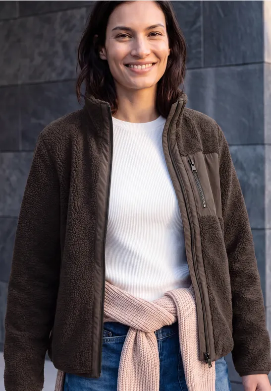 Eine lächelnde Frau trägt eine braune Fleecejacke, ein weißes Shirt und hat einen Pullover um die Hüfte gebunden.
