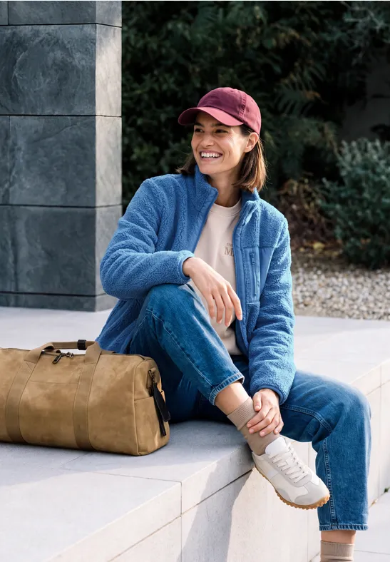 Eine Frau mit roter Mütze und blauer Fleecejacke sitzt lächelnd mit Tasche auf einer Steinbank.