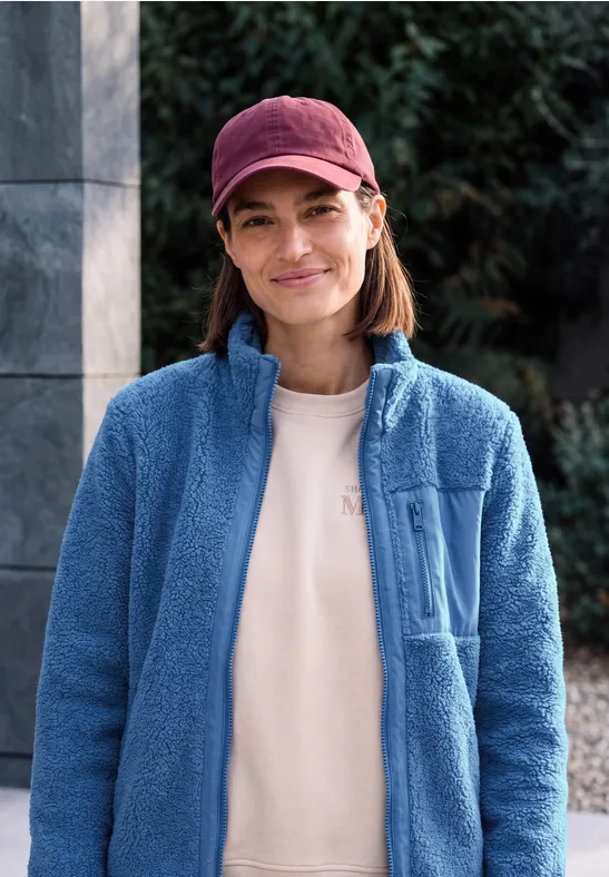 Frau mit roter Kappe und blauer Fleecejacke lächelt vor grünem Hintergrund.