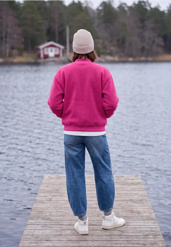 Person in pinkem Pullover und weißer Mütze steht auf Steg, blickt über ruhigen See mit Waldhaus.