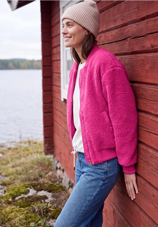Eine Frau in pinker Fleecejacke lehnt lächelnd an einer roten Holzwand am Ufer.