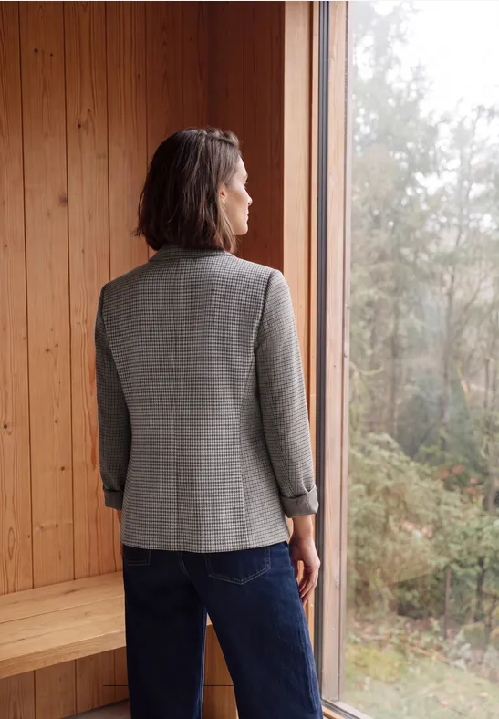 Vrouw in een grijze blazer en spijkerbroek, rug naar camera, kijkt door raam naar herfstig bos.
