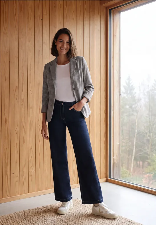 Een vrouw in jeans, wit topje, vest en sneakers staat voor een houten wand naast een groot raam.