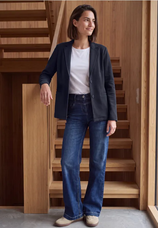 Une femme en jeans, t-shirt blanc et blazer bleu foncé sourit appuyée contre un escalier en bois.