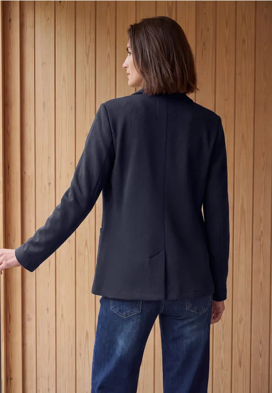 Une femme de dos portant un blazer noir et un jean bleu, devant un mur de lattes en bois.