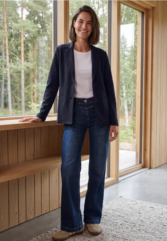 Femme en veste bleue et jean évasé appuyée sur une fenêtre en bois, forêt visible dehors.