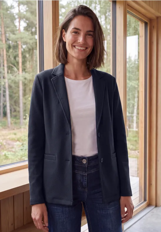 Une femme souriante en blazer et jean se tient devant une fenêtre offrant une vue sur la forêt.
