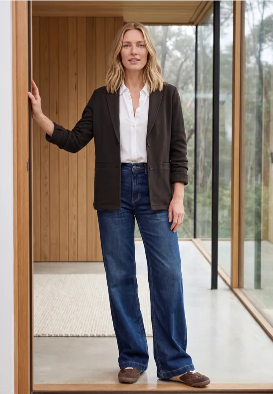 Une femme en chemise blanche, veste noire et jeans évasés se tient dans l’entrée d’une maison moderne et lumineuse.