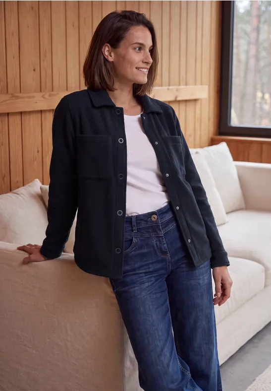 Femme appuyée sur un canapé, porte un jean, un t-shirt blanc et une surchemise noire dans une pièce en bois.