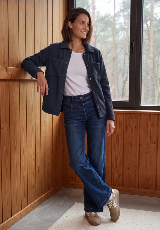 Femme en t-shirt blanc, veste et jean, appuyée et souriante contre un mur en bois près d’une fenêtre.