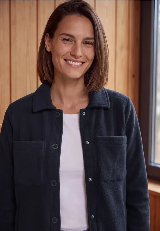 Une femme souriante porte un t-shirt blanc et une surchemise bleu foncé devant un mur en bois.
