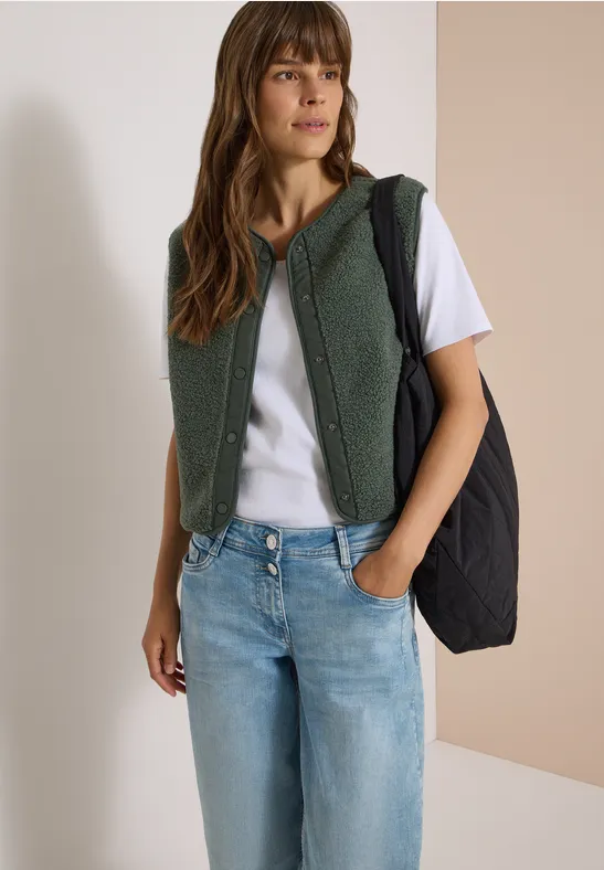 Femme portant un t-shirt blanc, un jean, une veste teddy verte à capuche et un sac noir.