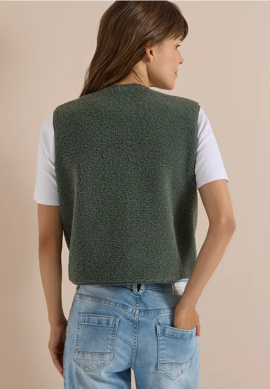 Une femme vue de dos portant un gilet en polaire vert gris, un T-shirt blanc et un jean.