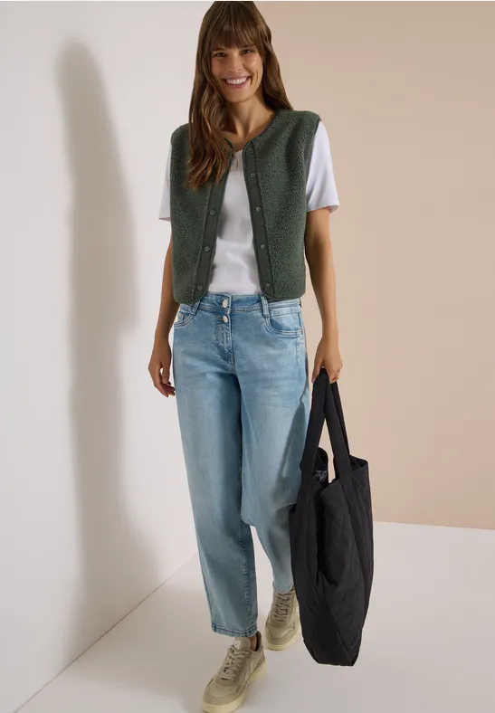 Femme en t-shirt blanc, gilet vert, jean bleu clair et baskets, souriante, portant un grand sac noir