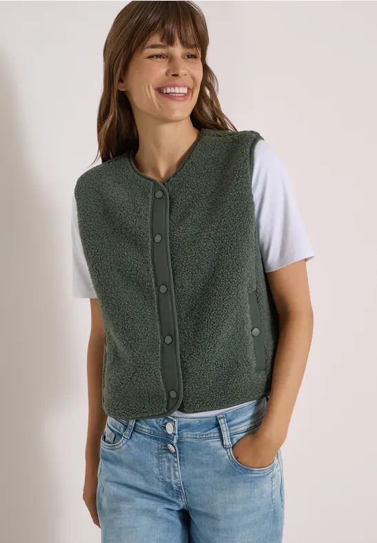 Une femme souriante porte une veste polaire verte à boutons sur t-shirt blanc et jean, une main en poche.