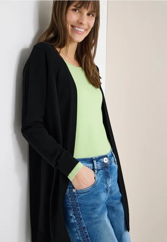 Frau trägt schwarzen Cardigan, grünes Top und Jeans, lehnt entspannt an weißer Wand.