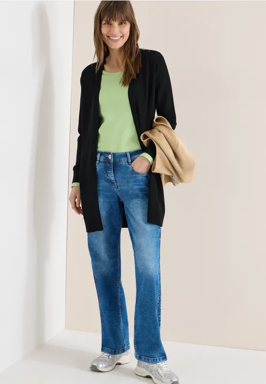 Modisches Outfit: schwarze Strickjacke, grünes Top, blaue Jeans, weiße Sneaker und beiger Mantel.