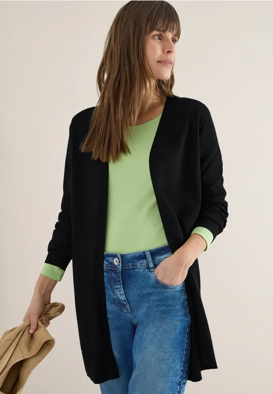 Frau trägt grünes Shirt, schwarze Strickjacke und Jeans, hält beigen Mantel.