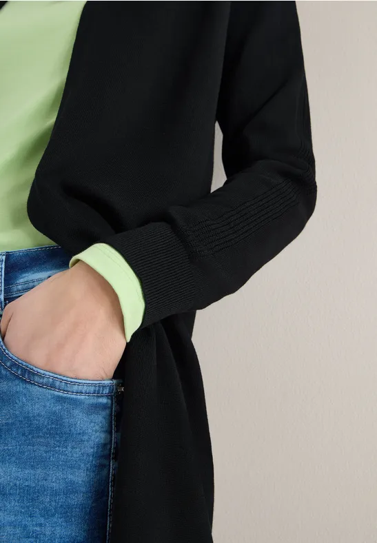 Person trägt schwarze Strickjacke, hellgrünes Top und blaue Jeans, Hand in Hosentasche.