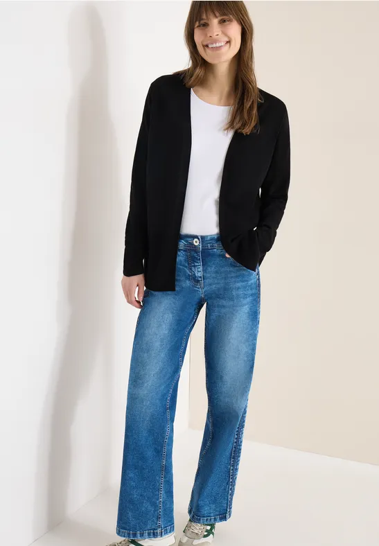 Frau trägt weißes T-Shirt, schwarzen Cardigan, weite blaue Jeans und weiße Sneaker, lehnt an einer Wand.