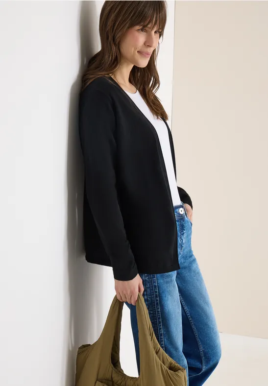 Frau lehnt an Wand, trägt schwarzen Cardigan, weißes Top, blaue Jeans und hält eine grüne Tasche.