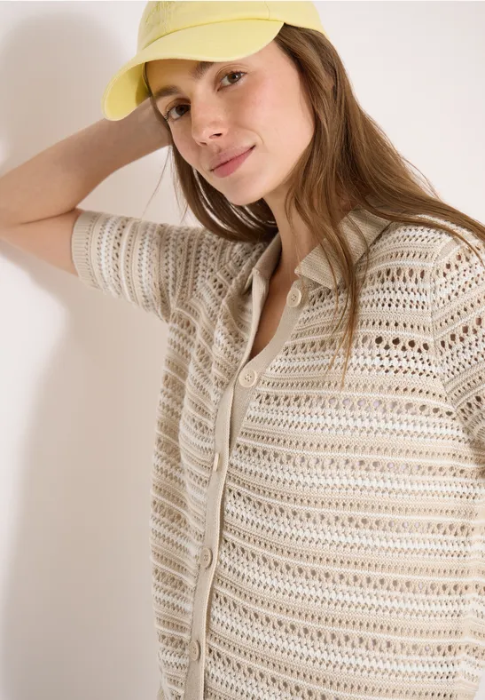 Une personne appuyée contre un mur porte un gilet en crochet beige clair et une casquette jaune.