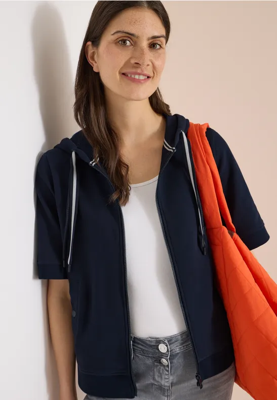 Person in dunkelblauer Kapuzenjacke über weißem Top trägt eine orangefarbene Tasche vor hellem Hintergrund.