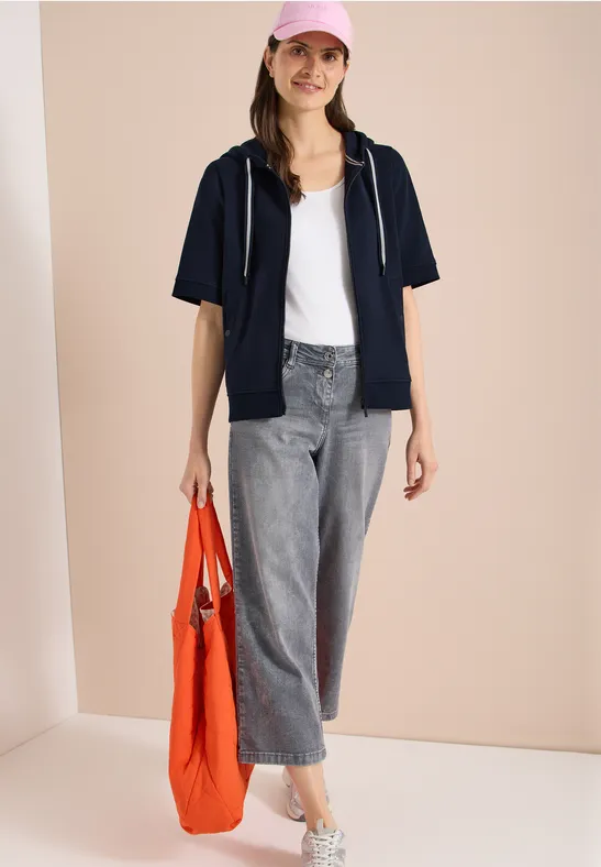 Frau mit weißem Top, dunkelblauer Kurzarmjacke, grauen Jeans und orangefarbener Tasche.