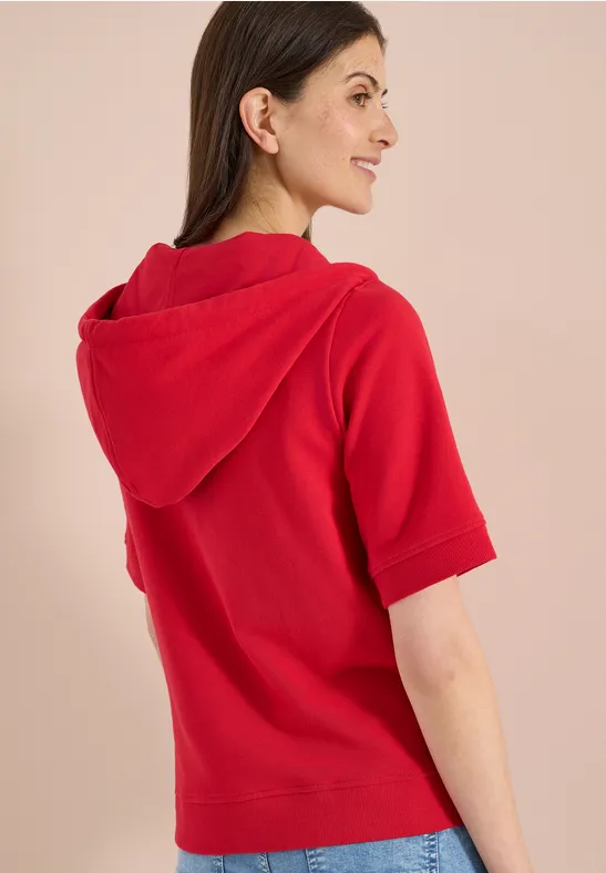 Rückansicht einer Frau in rotem kurzärmligem Kapuzenpullover vor beigem Hintergrund.