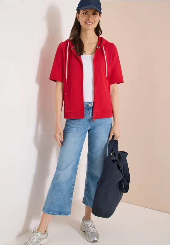 Frau in rotem Kapuzen-Shirt, weißem Top, weiter Jeans, silbernen Sneakern und großer Tasche.