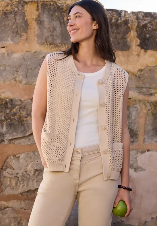 Une femme porte une veste en maille beige, un top blanc et tient une pomme verte devant un mur de pierre.