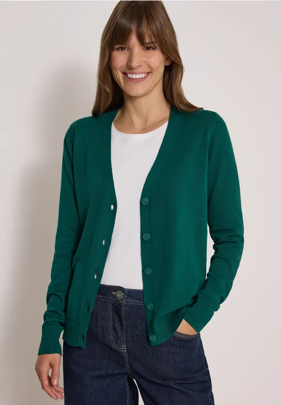 Eine lächelnde Frau trägt grünen Cardigan, weißes T-Shirt und blaue Jeans vor hellem Hintergrund.