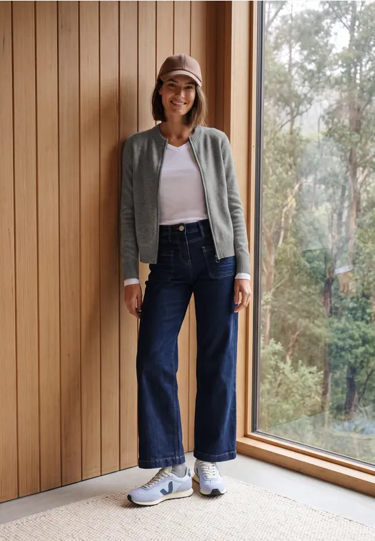 Frau in Baseballcap, grauem Cardigan, weißem Top und weiten Jeans steht neben Fenster und Holzpaneelen.